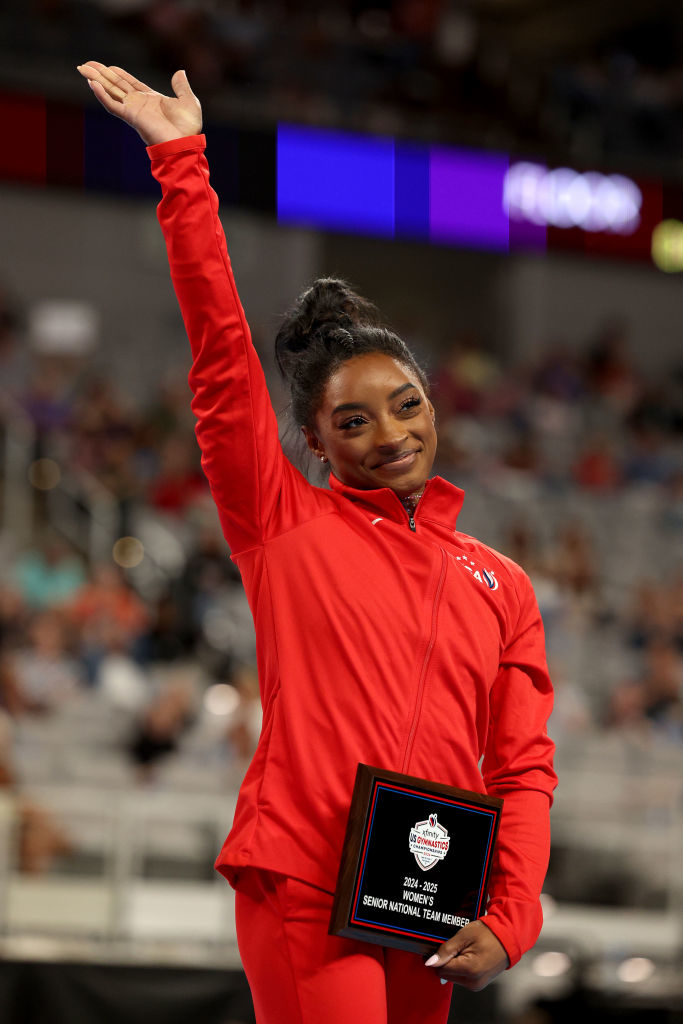 Simone Biles waves while holding a plaque at a gymnastics event, wearing an athletic tracksuit
