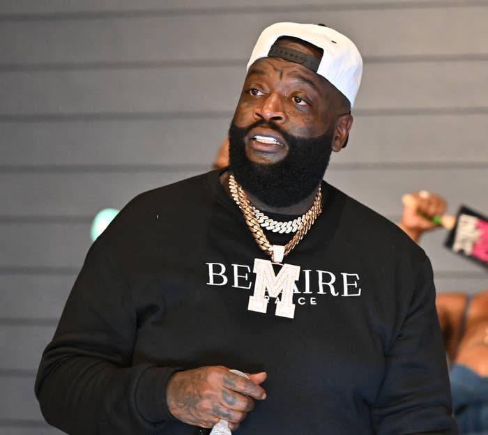 Rick Ross wears a black sweater with &quot;BE MIRE&quot; text, layered chains, and a white cap, appearing at a music event, with another person blurred in the background