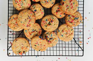 Cookies with colorful sprinkles rest on a cooling rack, with some sprinkles scattered on a white surface below