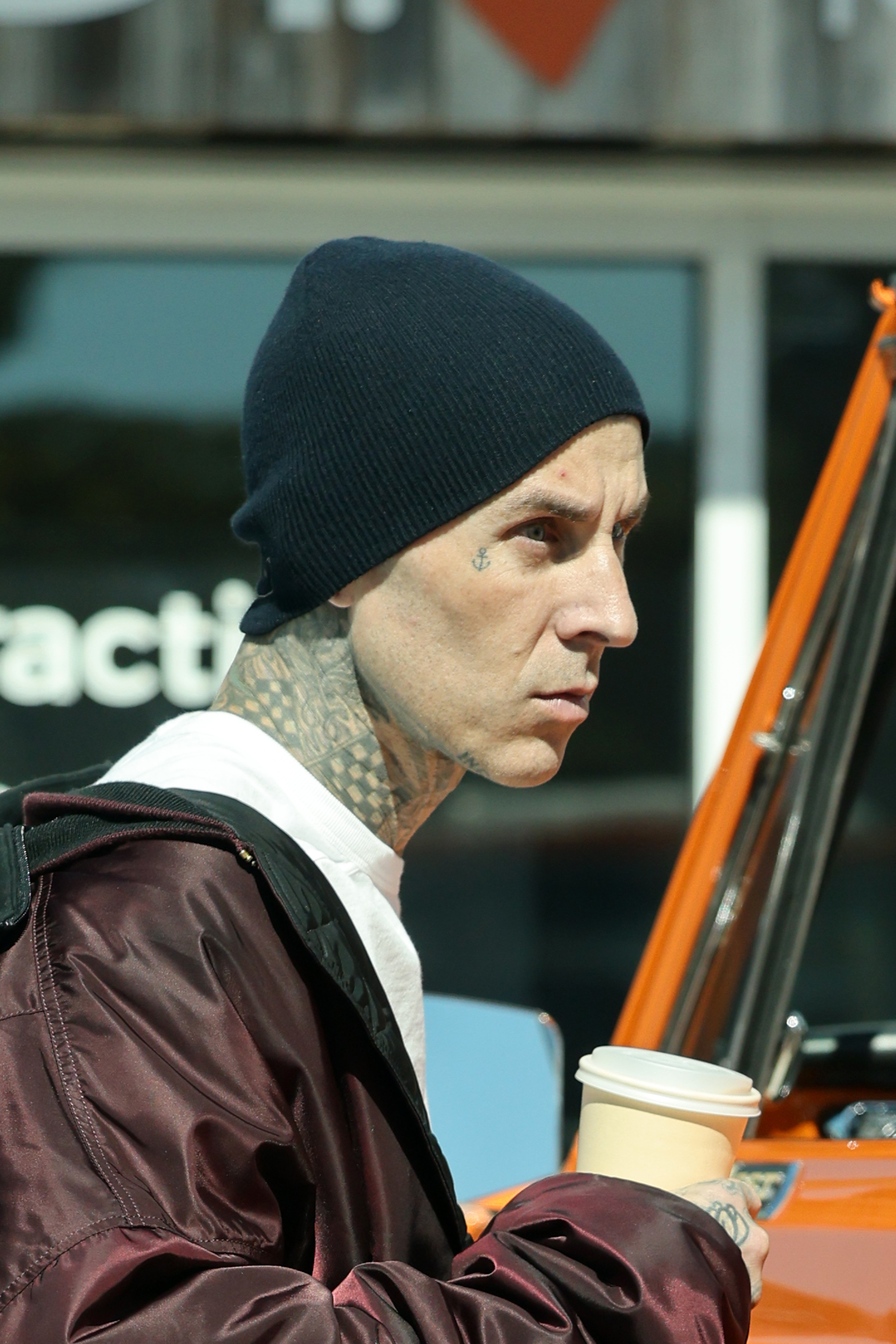 Travis Barker is seen outdoors, wearing a beanie, jacket, and holding a coffee cup. Part of his neck tattoos are visible