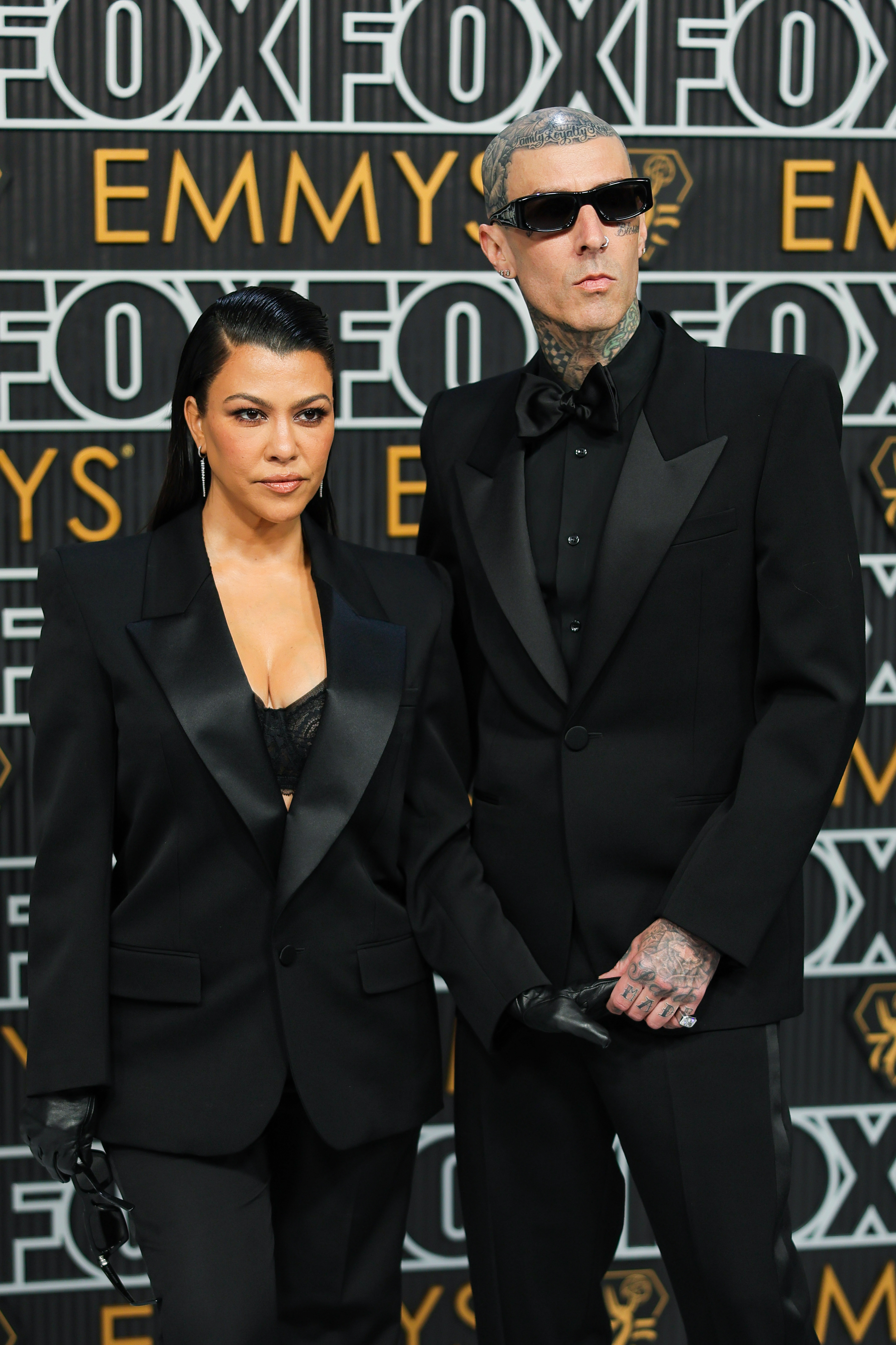 Kourtney Kardashian and Travis Barker at the Emmys red carpet, both wearing matching black suits with black shirts. Travis has tattoos and wears sunglasses