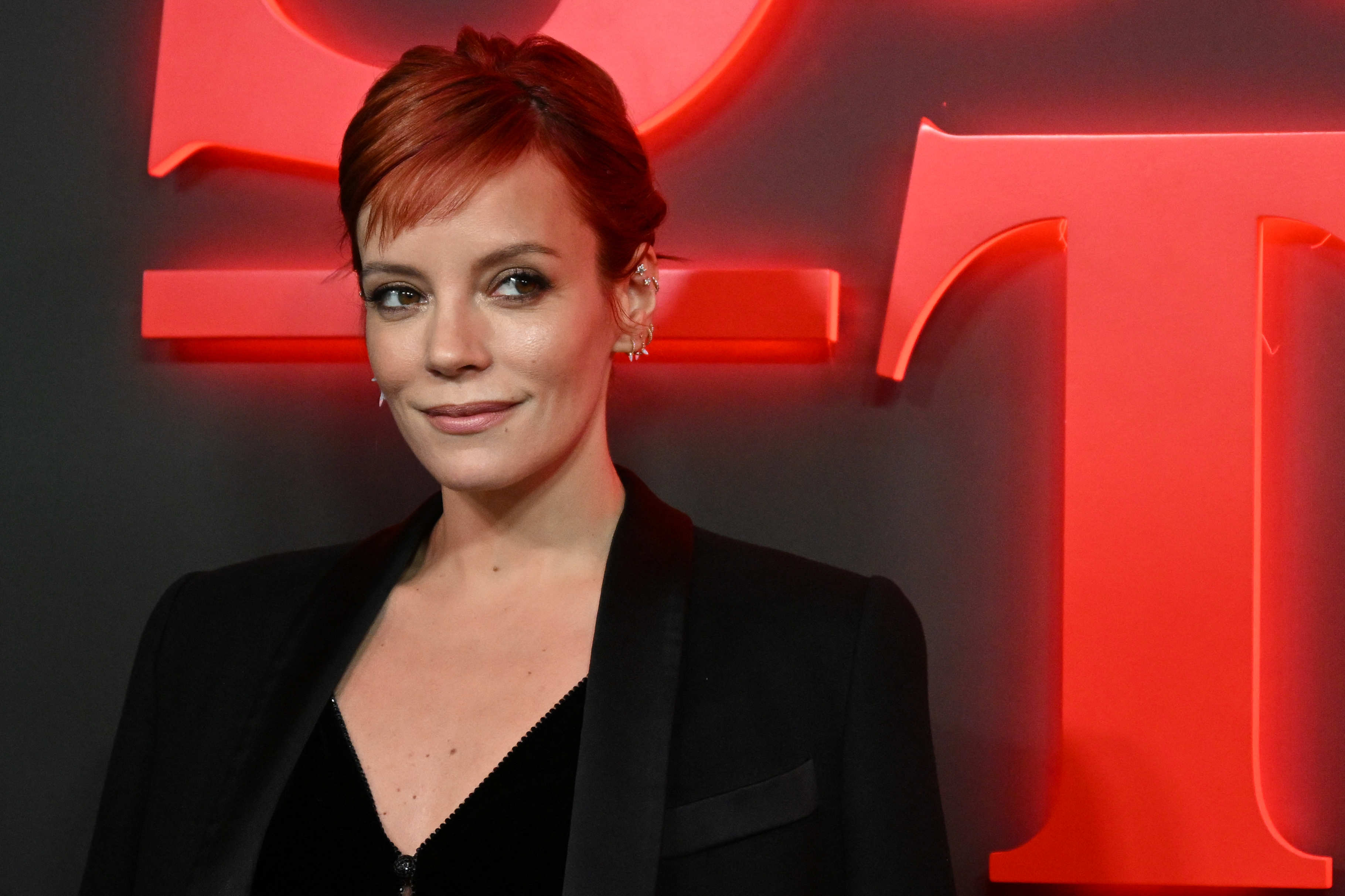 Lily Allen smiles while wearing a dark blazer in front of a neon background