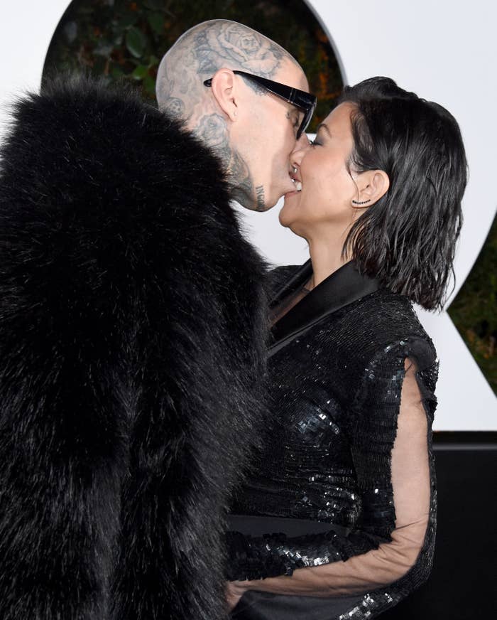Travis Barker and Kourtney Kardashian share a kiss; Barker in a black fur coat and oversized sunglasses, Kardashian in a black sequined dress