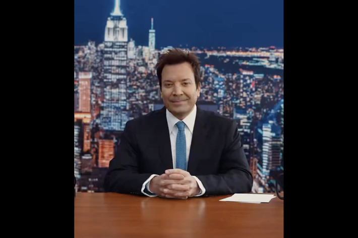 Jimmy Fallon sits at a desk in front of a cityscape background, smiling and wearing a black suit and tie