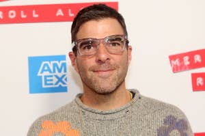 Zachary Quinto wearing a floral patterned sweater and glasses at an event with AMEX branding in the background