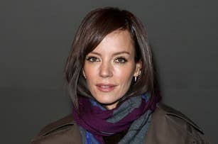 Lily Allen wearing a dark coat and a colorful scarf, holding a white fluffy bag. She has short, styled hair and is posing for the camera