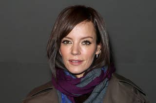 Lily Allen wearing a dark coat and a colorful scarf, holding a white fluffy bag. She has short, styled hair and is posing for the camera