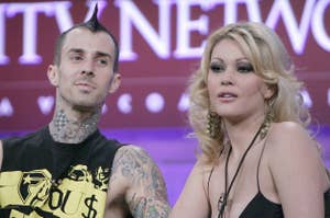 Travis Barker and Shanna Moakler appear on stage. Travis wears a sleeveless shirt showcasing his tattoos, while Shanna has long, wavy hair and dangles earrings