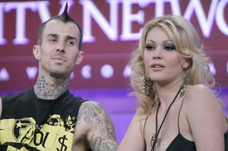 Travis Barker and Shanna Moakler appear on stage. Travis wears a sleeveless shirt showcasing his tattoos, while Shanna has long, wavy hair and dangles earrings