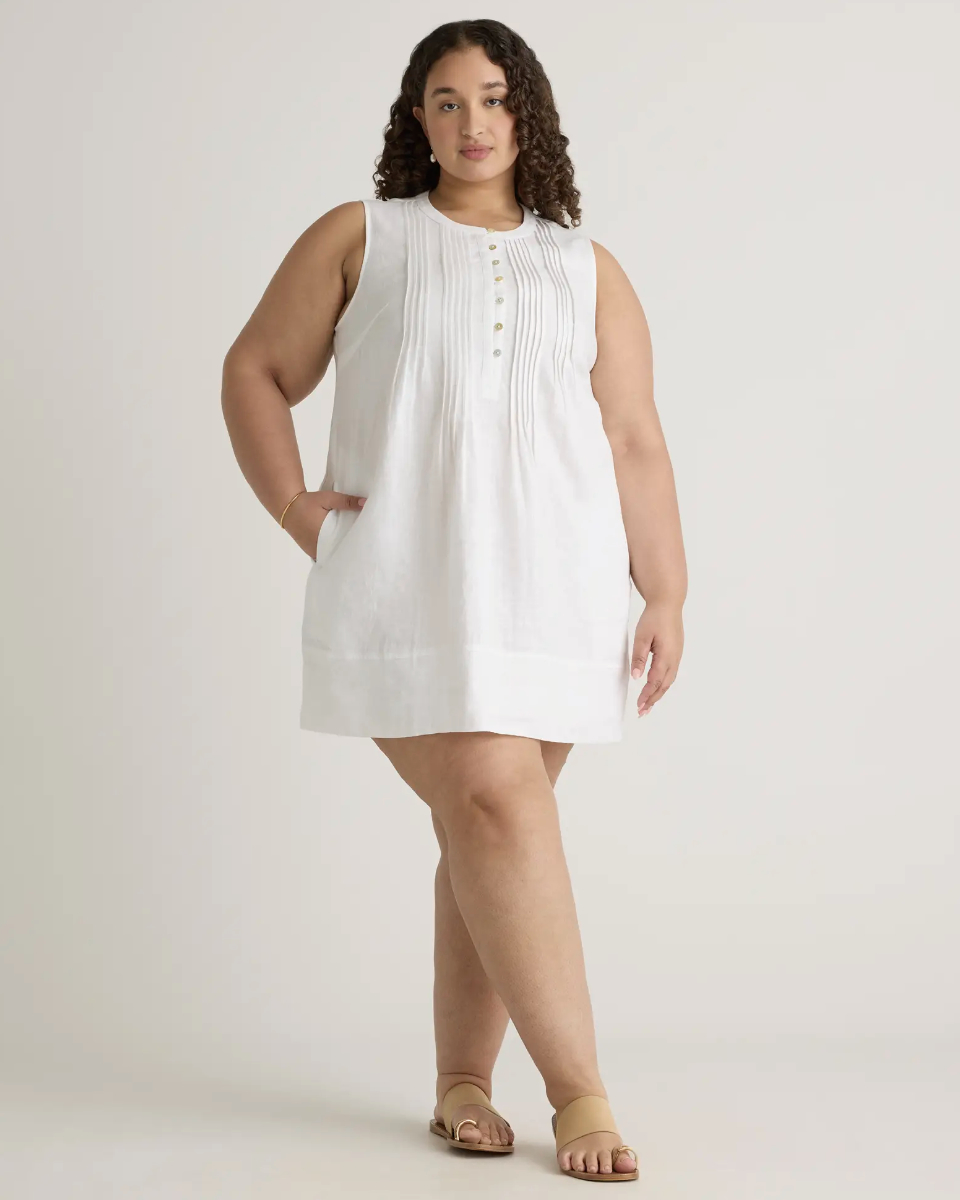 A Model wearing a sleeveless, knee-length linen dress with button detailing and sandals