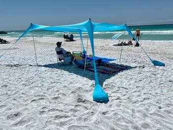 Orang duduk di bawah kanopi pantai biru di pantai berpasir. Di dekatnya adalah handuk pantai yang penuh warna dan perlengkapan pantai. Lautan ada di latar belakang