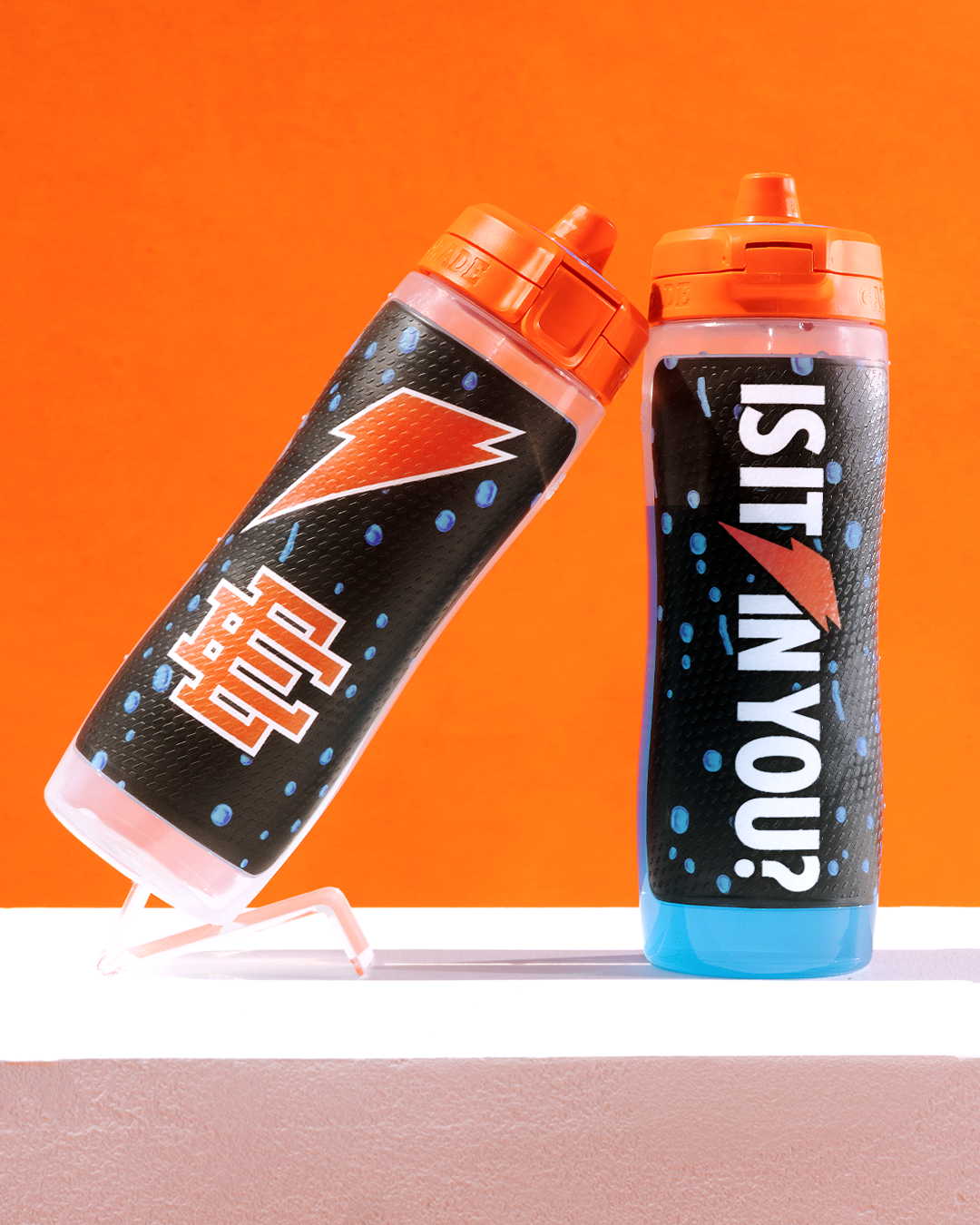Two sports drink bottles with blue liquid, orange caps, and lightning bolt designs are displayed. The bottle on the left features &quot;40&quot; and the one on the right reads &quot;Is it in you?&quot;