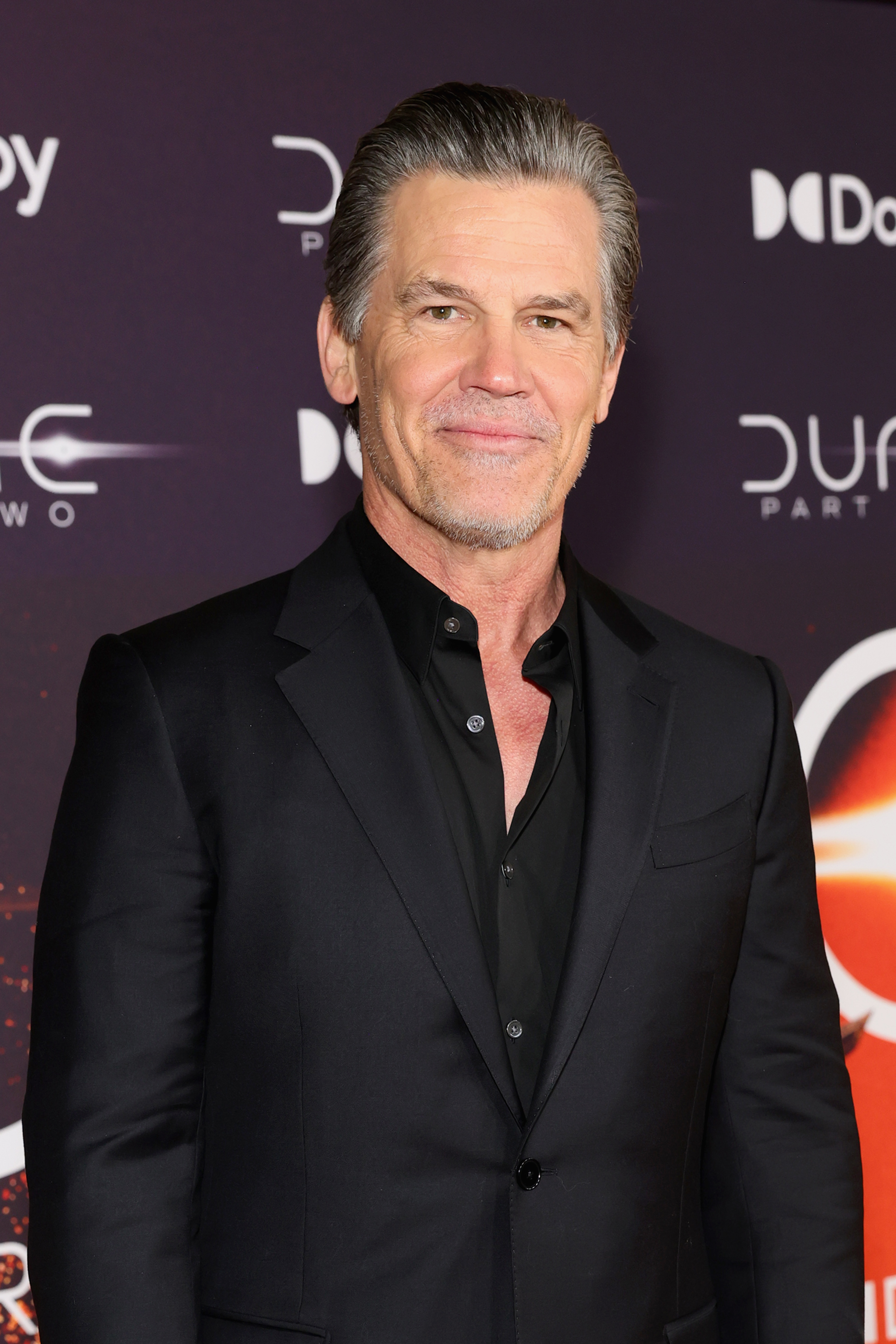 Josh Brolin on a red carpet, wearing a black suit with a black dress shirt, attending an event for "Dune: Part Two."
