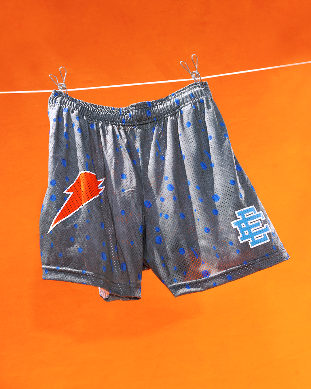 Gray athletic shorts with blue dots, featuring a red lightning bolt logo and a white &quot;BE&quot; monogram, hanging on a clothesline against an orange background