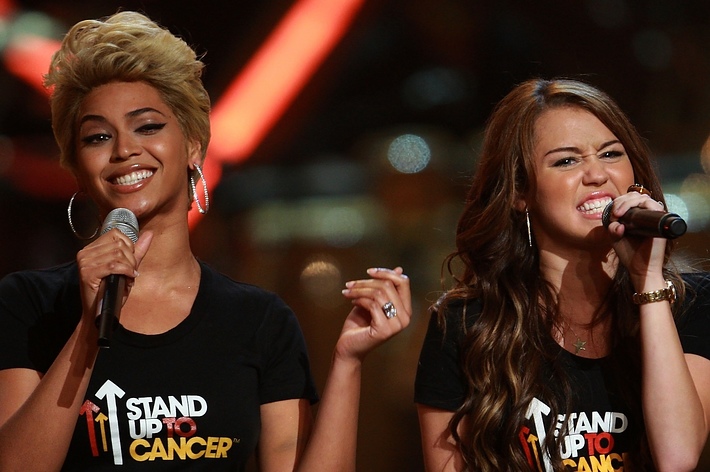 Beyoncé and Miley Cyrus perform at a Stand Up to Cancer event, both wearing black Stand Up to Cancer T-shirts