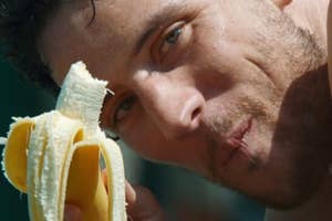 A person eating a banana while looking at the camera, close-up shot. The person's facial features are prominent, and they are taking a bite of the banana
