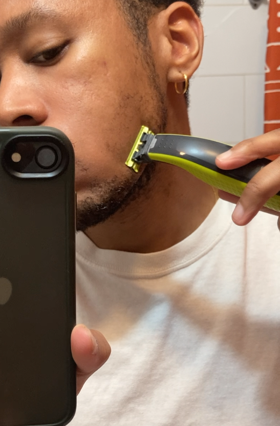 A person in a white shirt holds a phone and shaves their beard with an electric razor