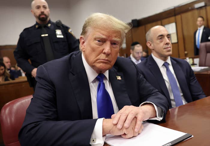 Donald Trump sitting at a table with folded hands, flanked by a police officer and his lawyer inside a courtroom