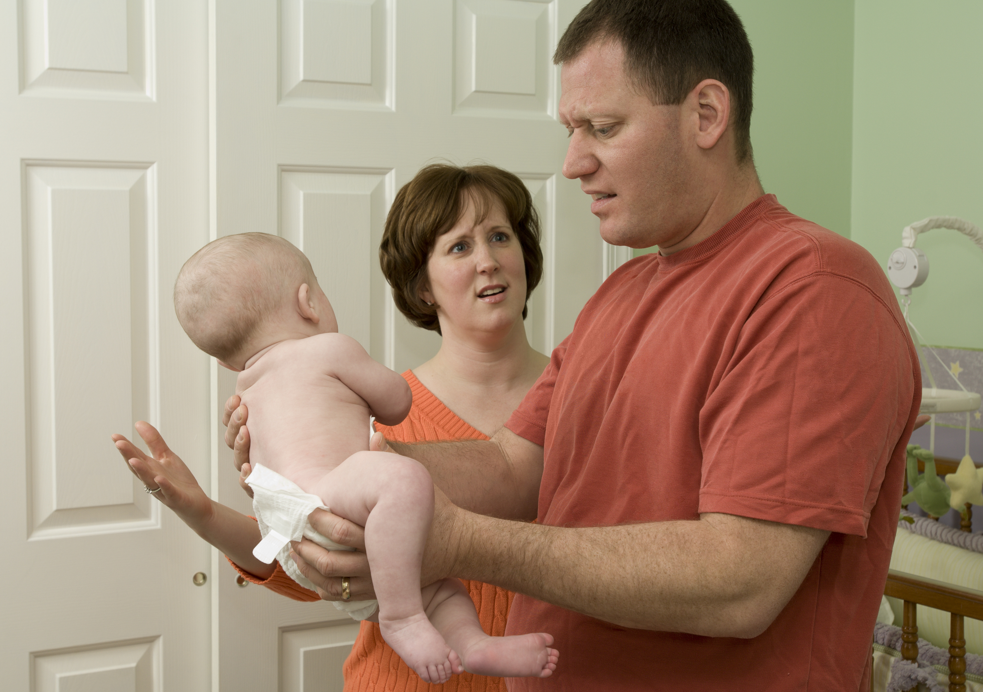 A man in a red shirt holds a baby with a distressed expression while a woman looks on in confusion. The scene appears to be in a nursery setting