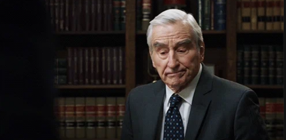 An older man with white hair wearing a dark suit and tie stands in front of bookshelves filled with books