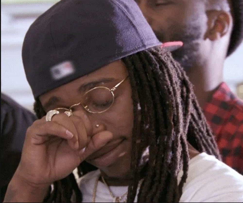 A man with dreadlocks wearing a cap and glasses wipes his eyes. Another man, also with dreadlocks, stands behind him with eyes closed