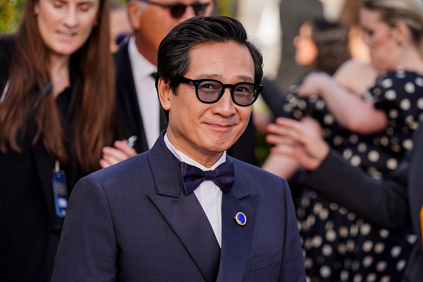Ke Huy Quan poses on the red carpet wearing a stylish dark suit with a bow tie and glasses, while attendees and photographers are visible in the background