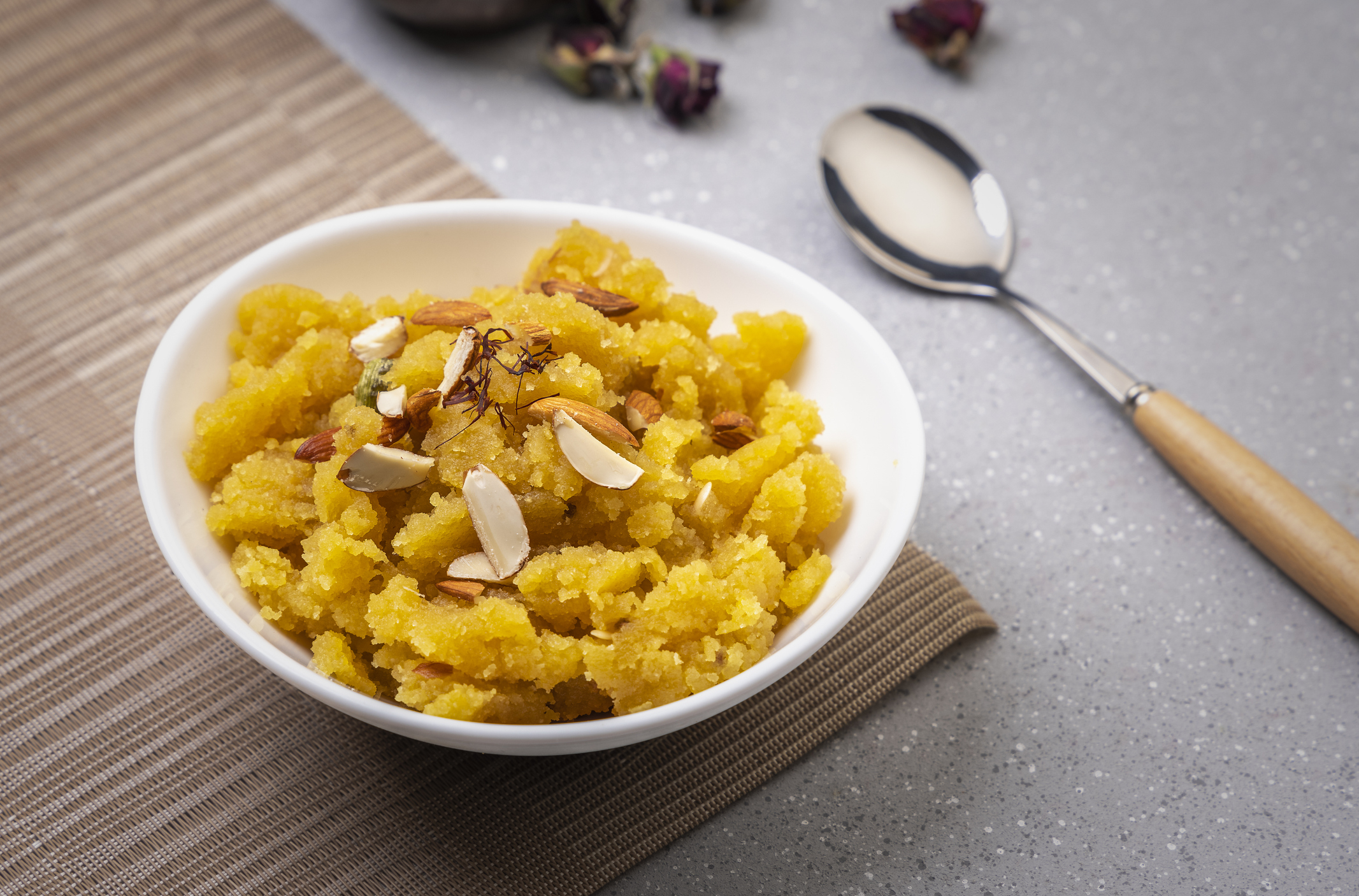 A bowl of halwa topped with sliced almonds and saffron, placed next to a spoon on a table