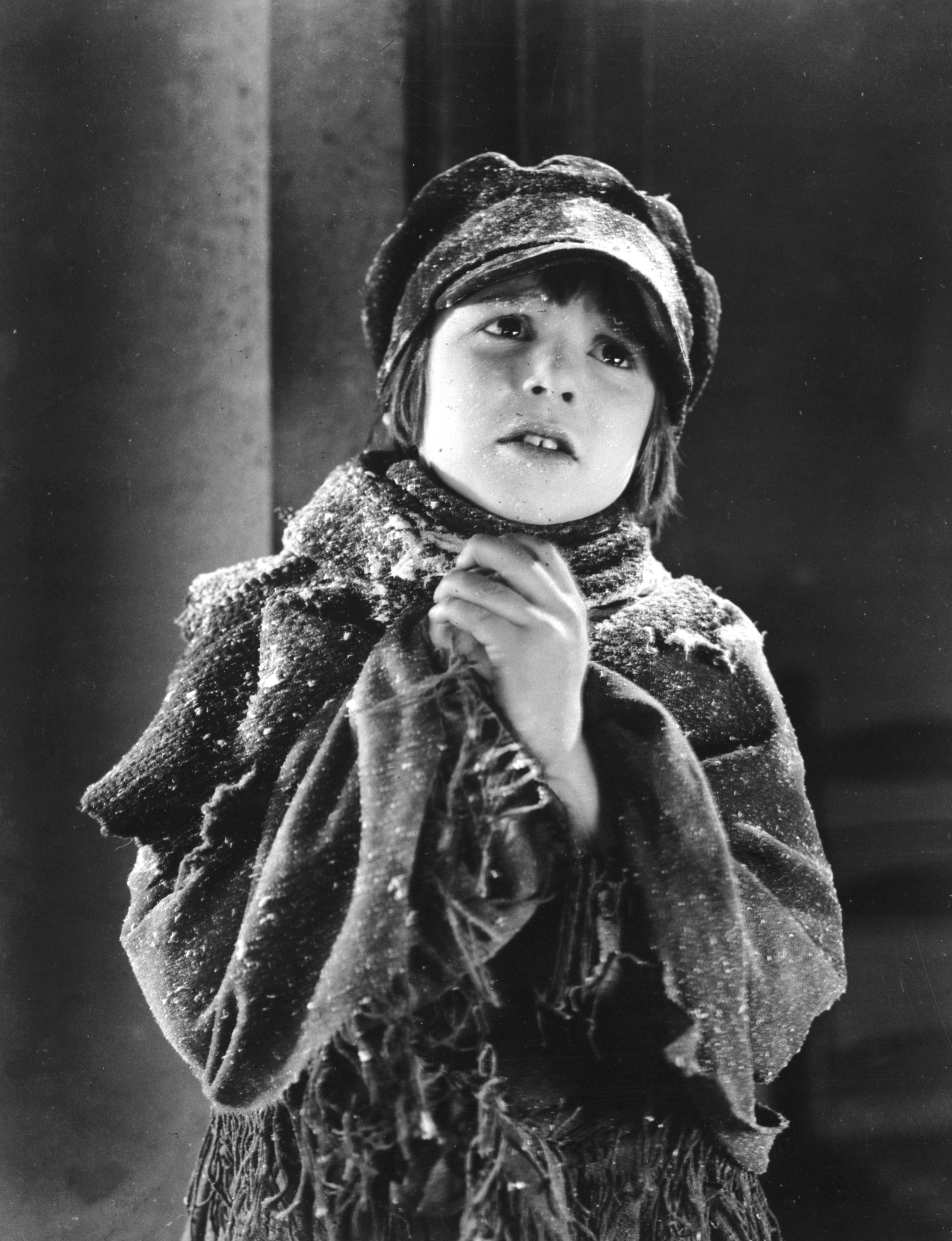 A child actor dressed in tattered clothing, wearing a cap and scarf, looks up with a hopeful expression