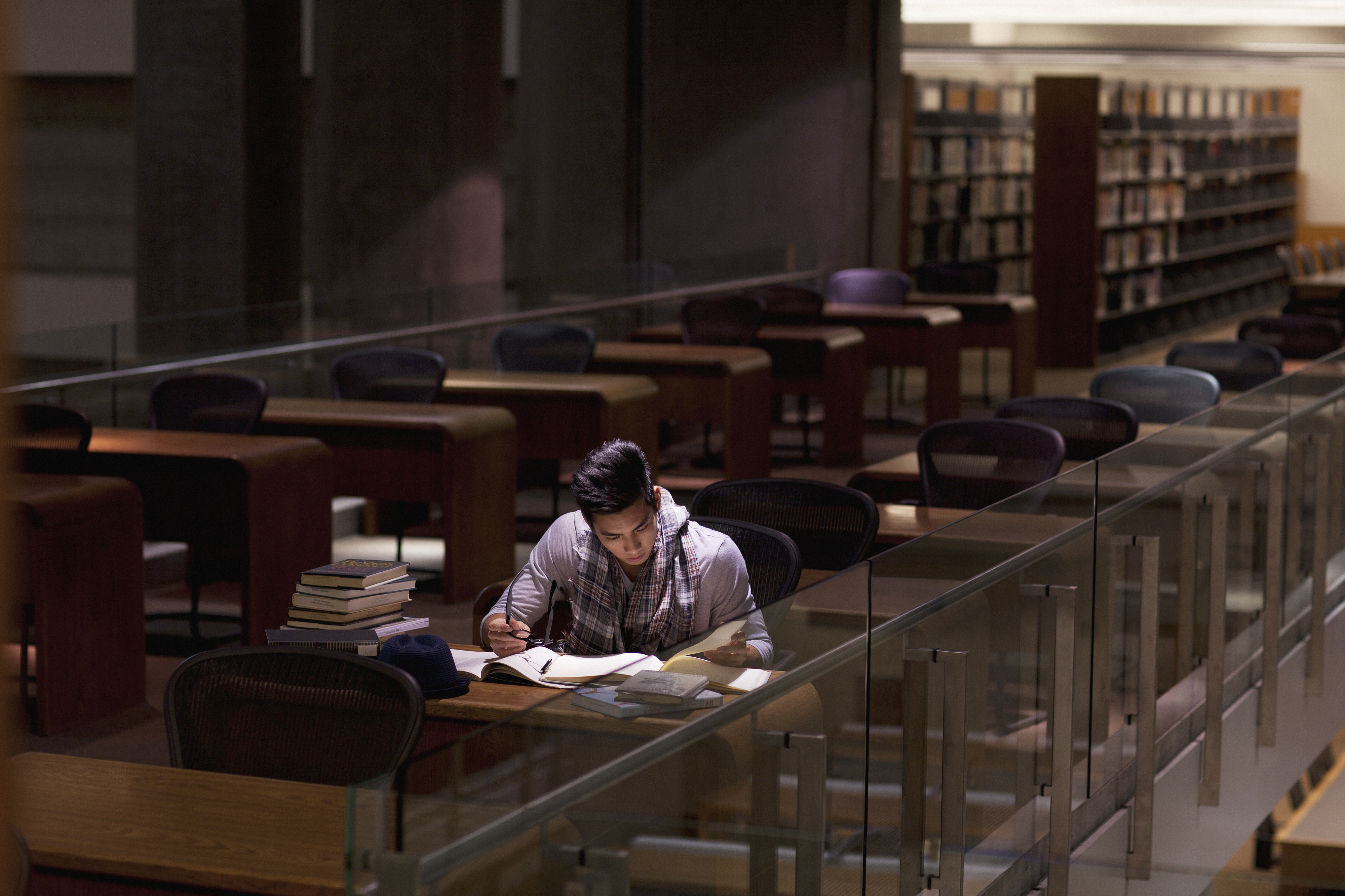 A person reads a book at a desk in a quiet library surrounded by empty chairs and shelves filled with books
