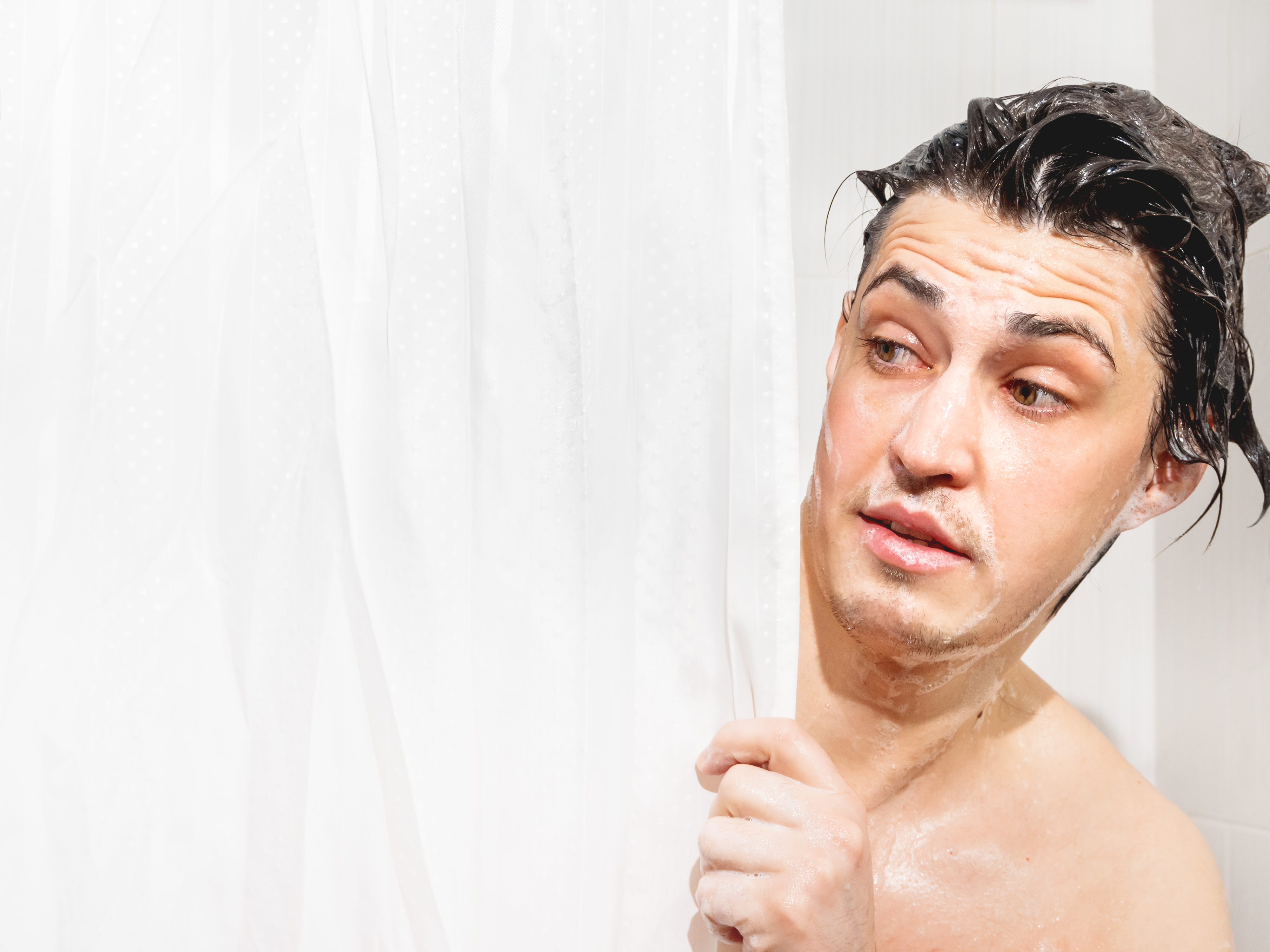 A person peeks out from behind a shower curtain, displaying a curious or cautious expression