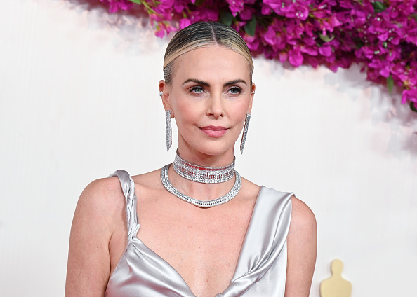 Charlize Theron on a red carpet wearing a silver dress with a plunging neckline, diamond choker necklace, and long earrings, standing in front of pink flowers