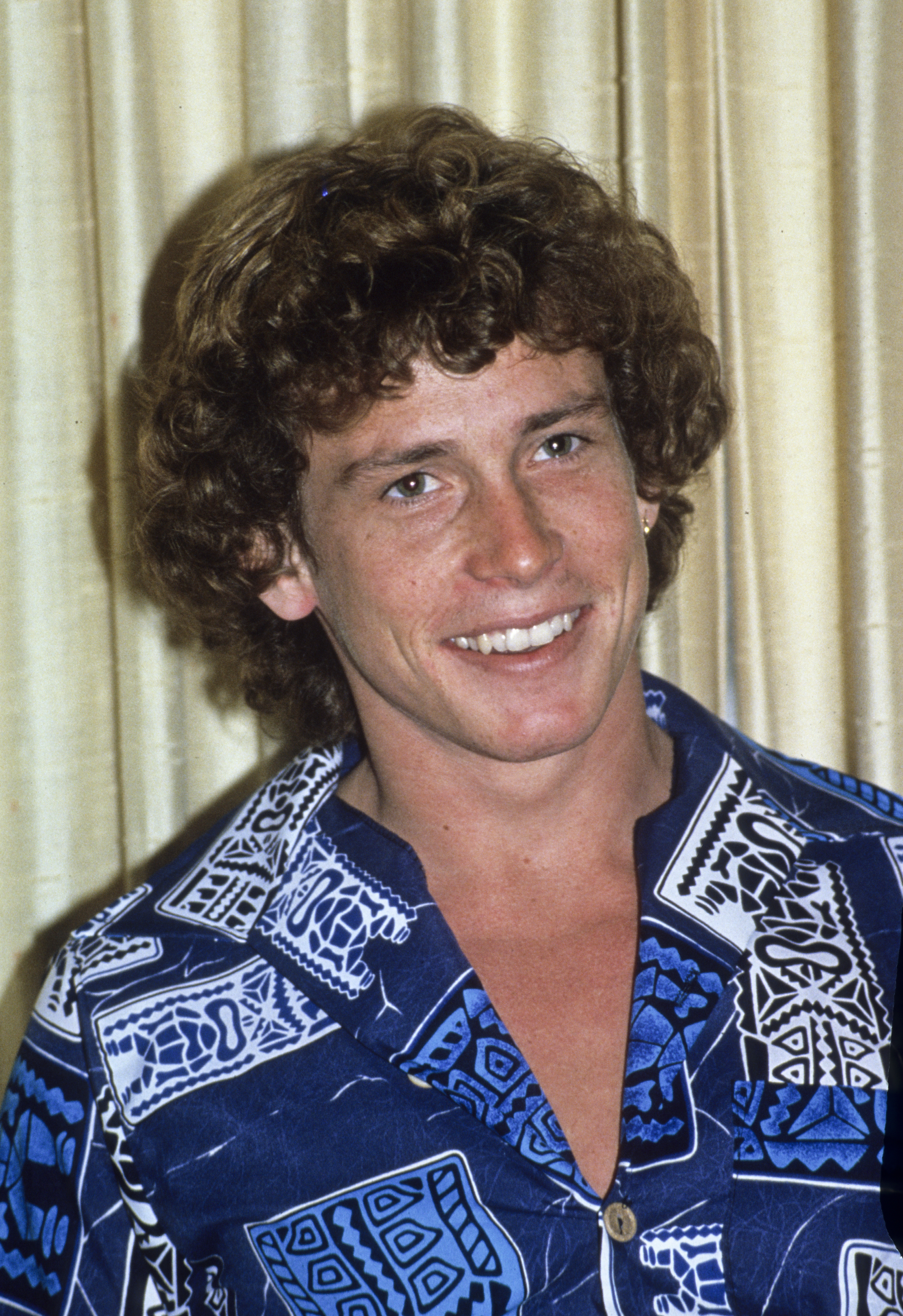 Smiling man with curly hair wearing a blue patterned shirt, standing in front of beige curtains
