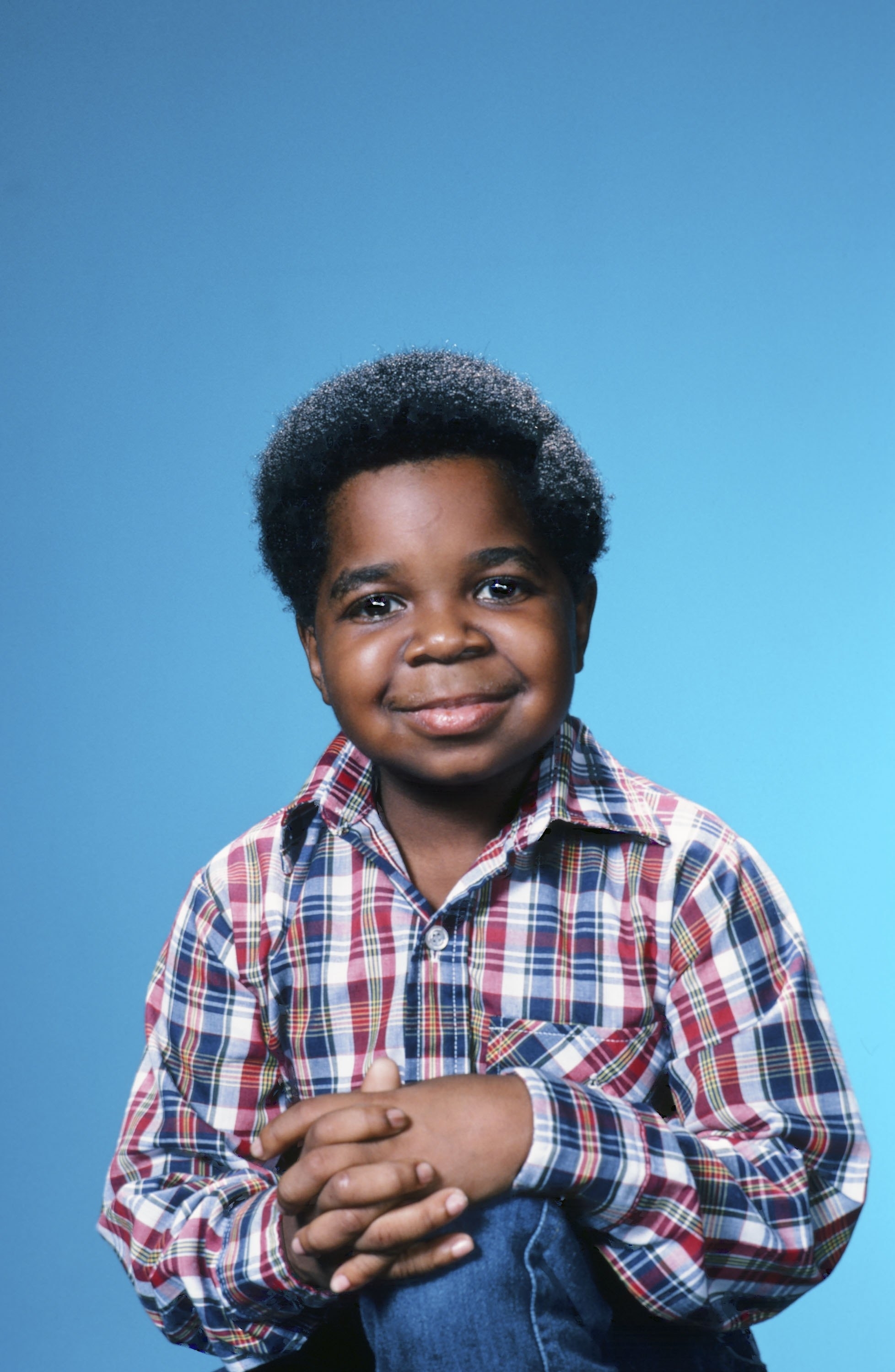 Gary Coleman is smiling, seated, and wearing a plaid shirt against a blue background