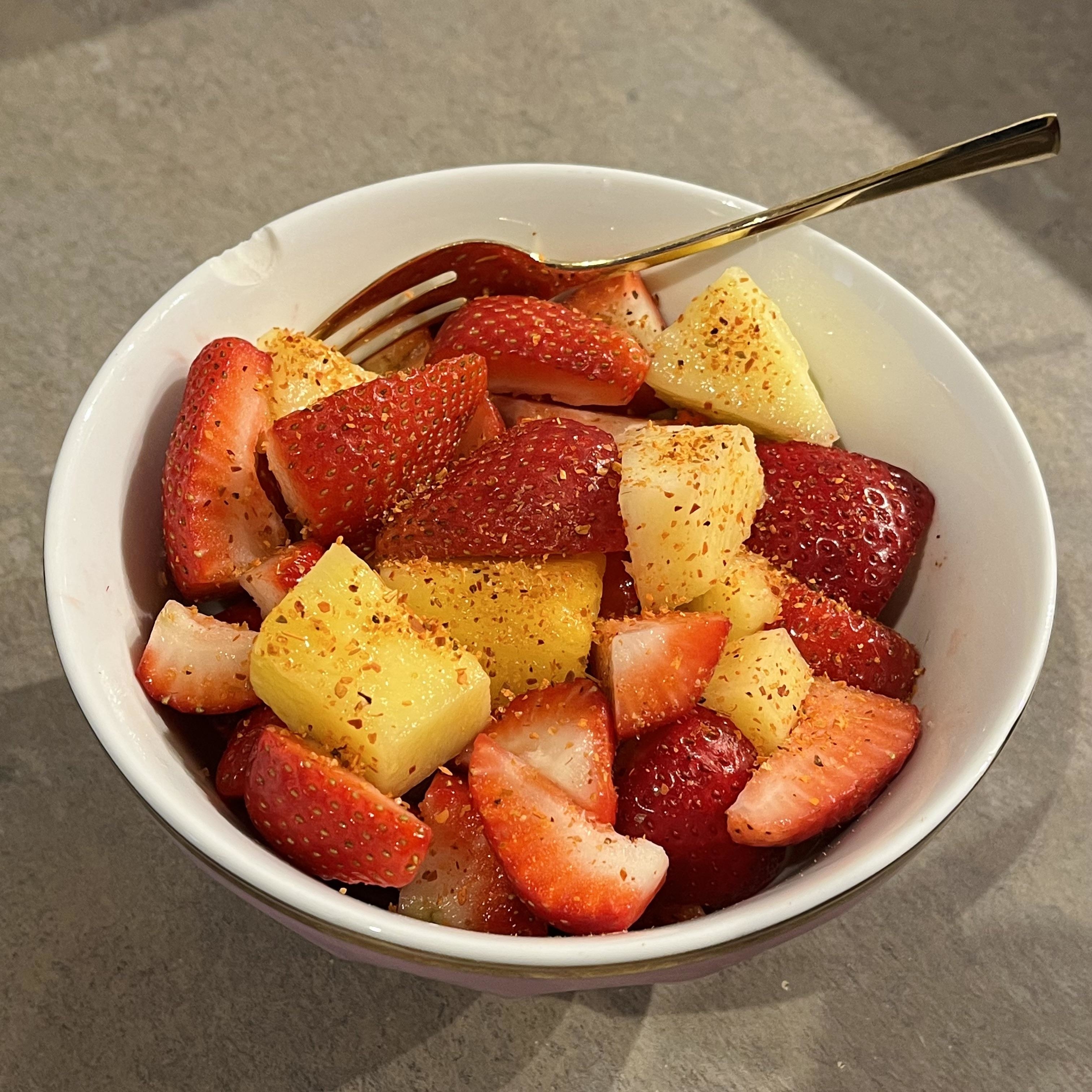 A bowl of cut strawberries and pineapple chunks, with chili powder sprinkled on top, and a small golden fork placed in the bowl