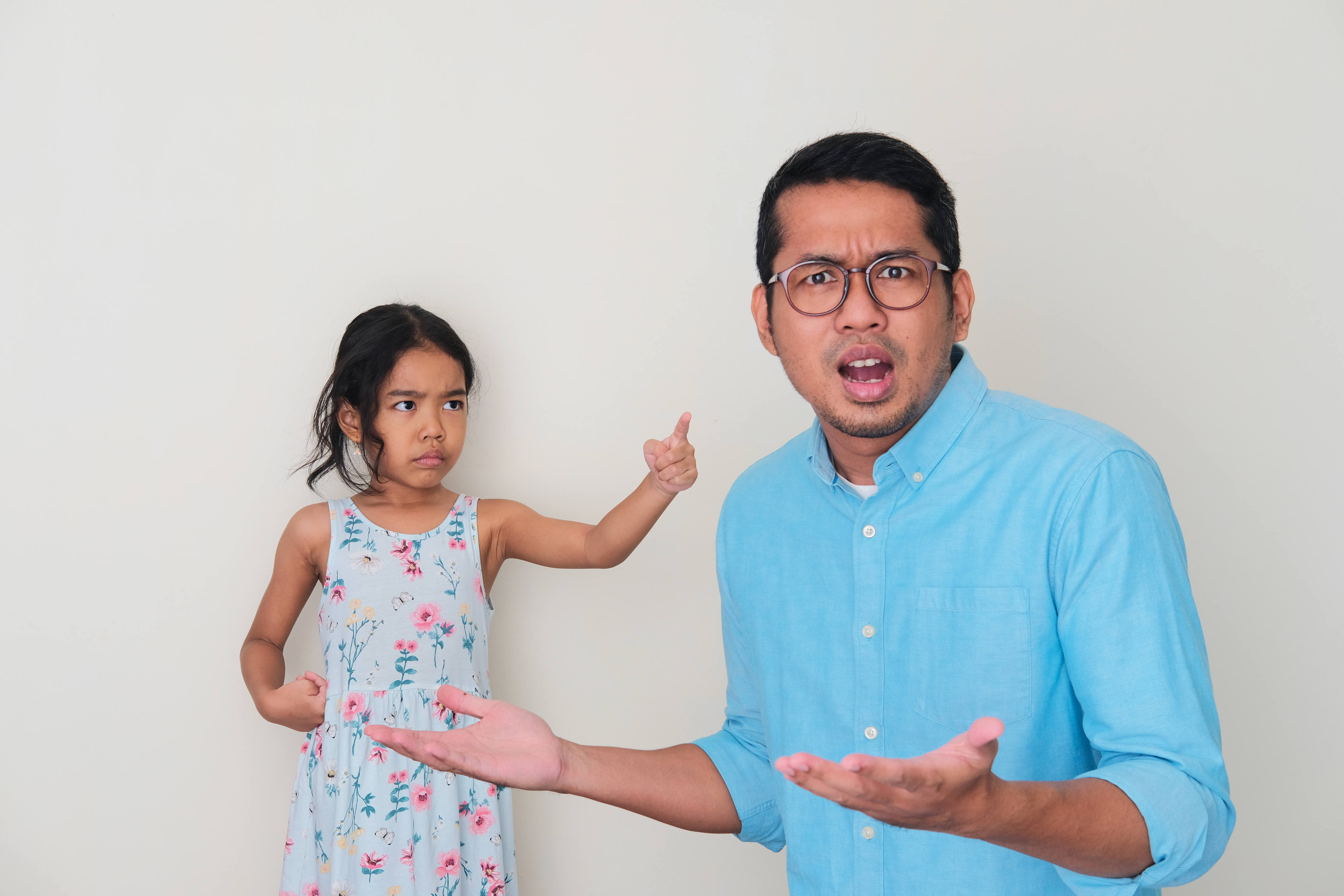 A man in a light blue shirt looks confused with his arms outstretched while a young girl in a floral dress points at him with a stern expression