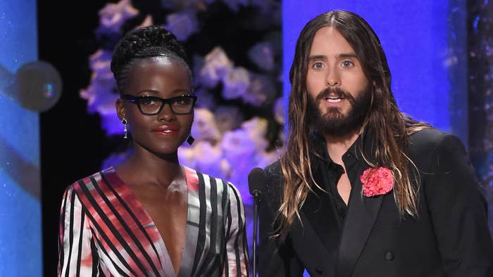 Lupita Nyong&#x27;o in a stylish striped dress and Jared Leto in a black suit with a pink rose lapel pin, both speaking on stage