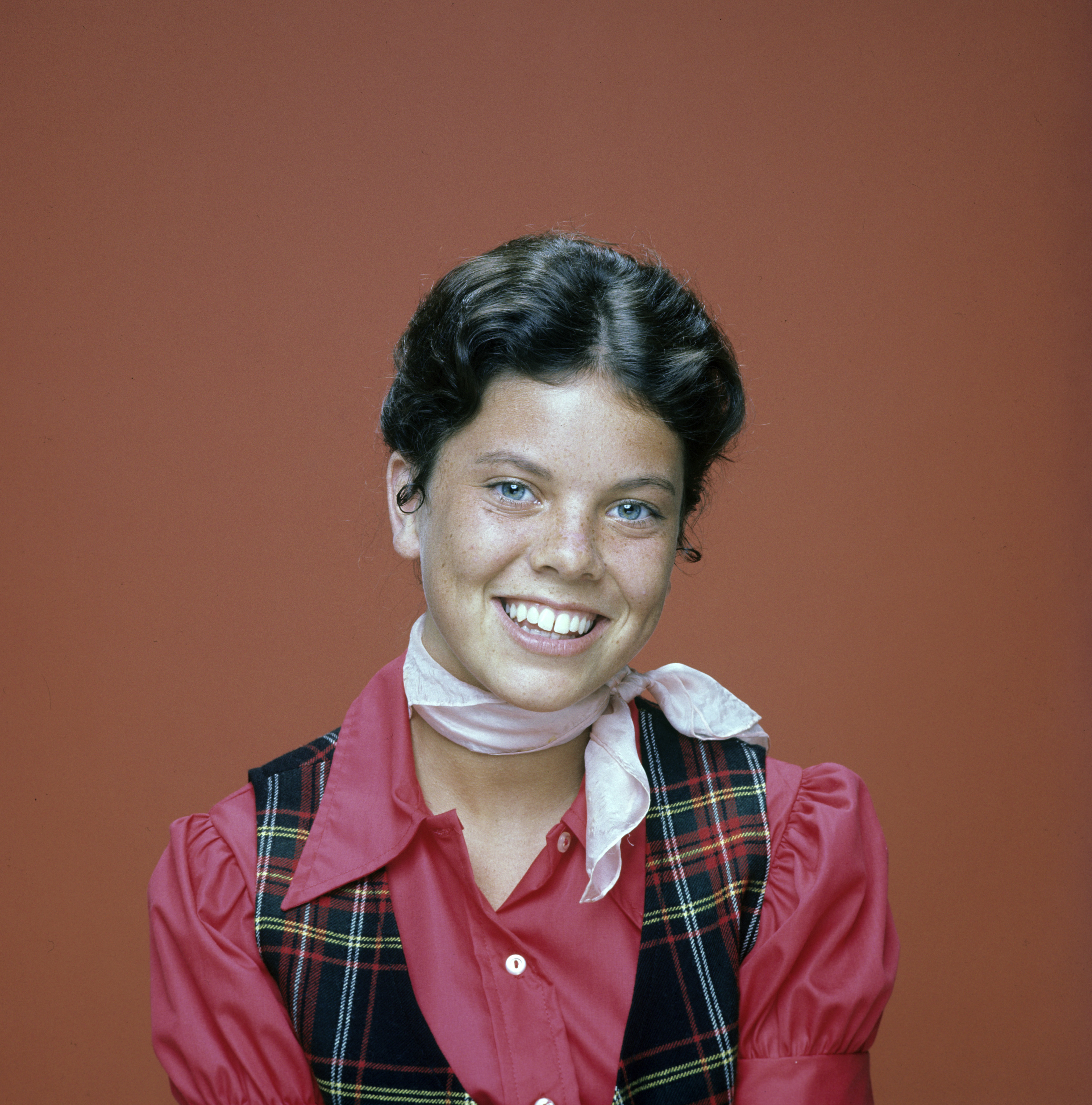 A smiling young woman with short dark hair wears a plaid vest over a collared shirt with a light scarf around her neck against a plain background