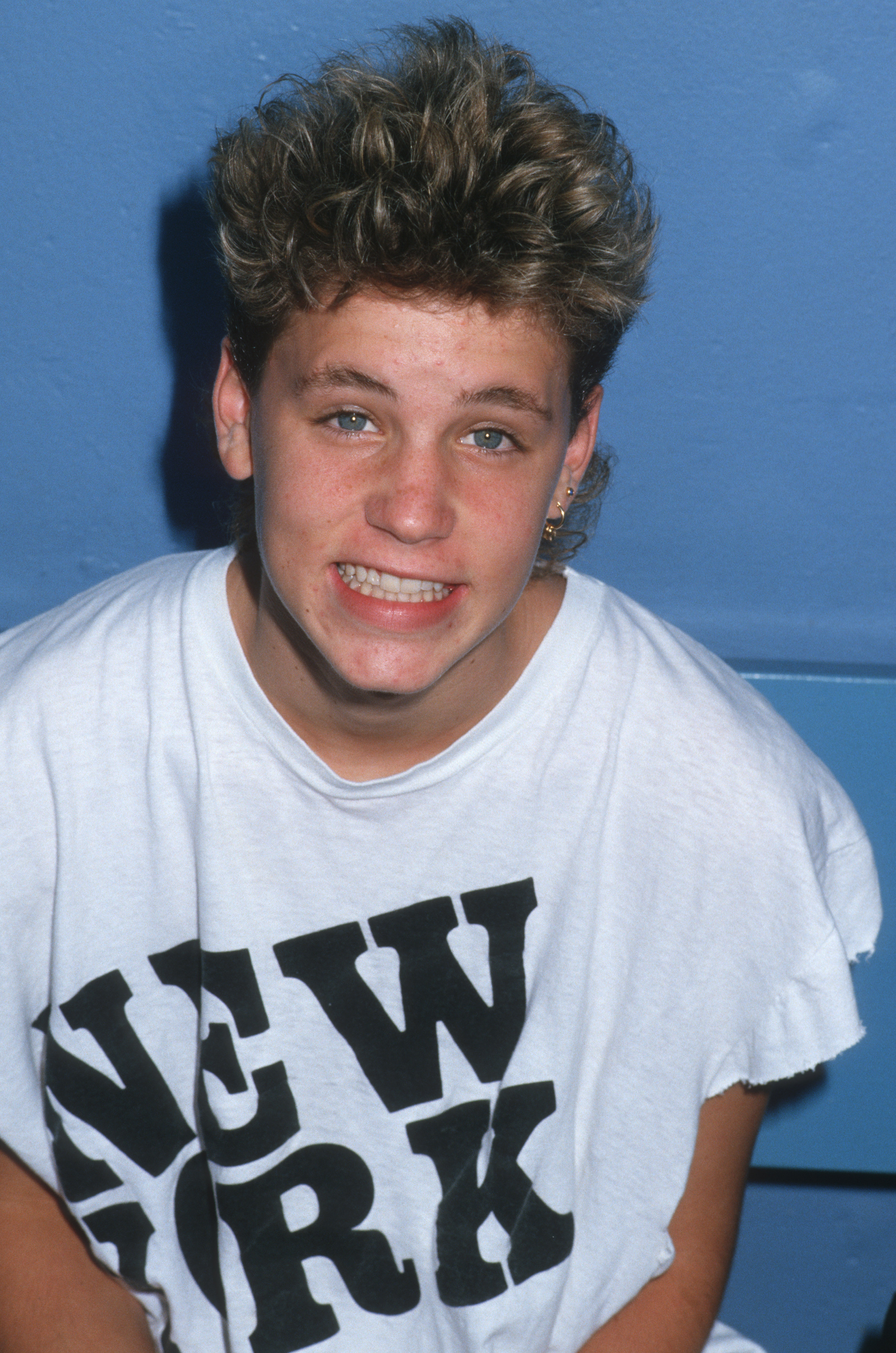 A person with short, curly hair and a slight smile wears a T-shirt that says &quot;NEW YORK.&quot; They are posing against a plain background