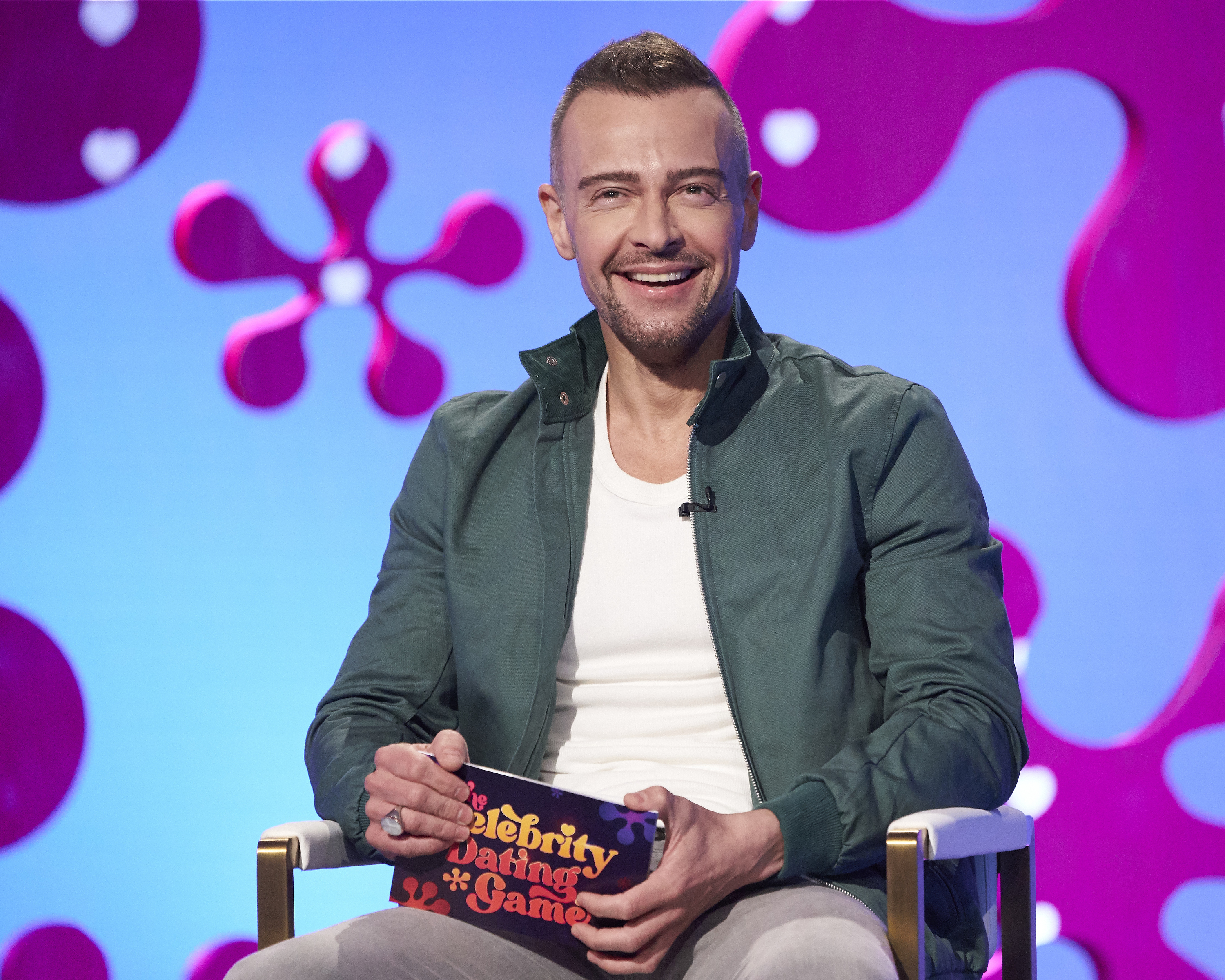Man seated and smiling on a game show set, holding a colorful sign that reads &quot;Celebrity Dating Game.&quot; He is wearing a white shirt and a green jacket