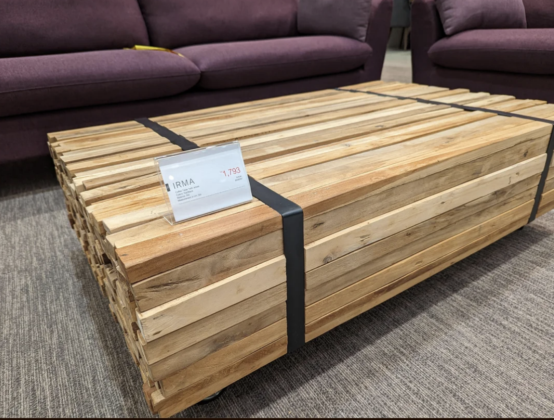 Wooden table with multiple slats and black straps on display in a furniture store. A price tag shows "IRMA 1,799." Two purple sofas are in the background