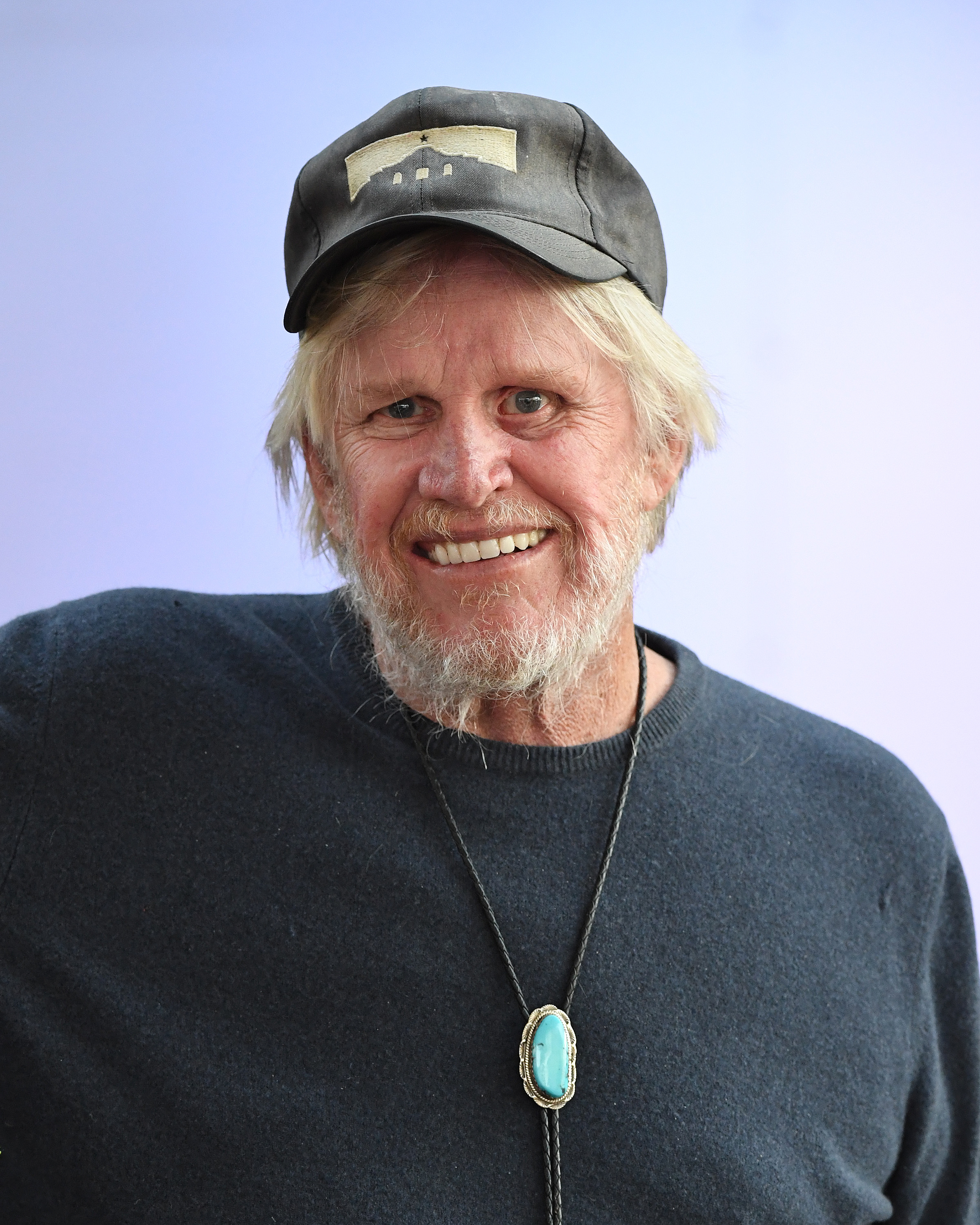 Older man with beard and baseball cap, Gary Busey, smiles and poses in a dark sweater with large turquoise pendant necklace