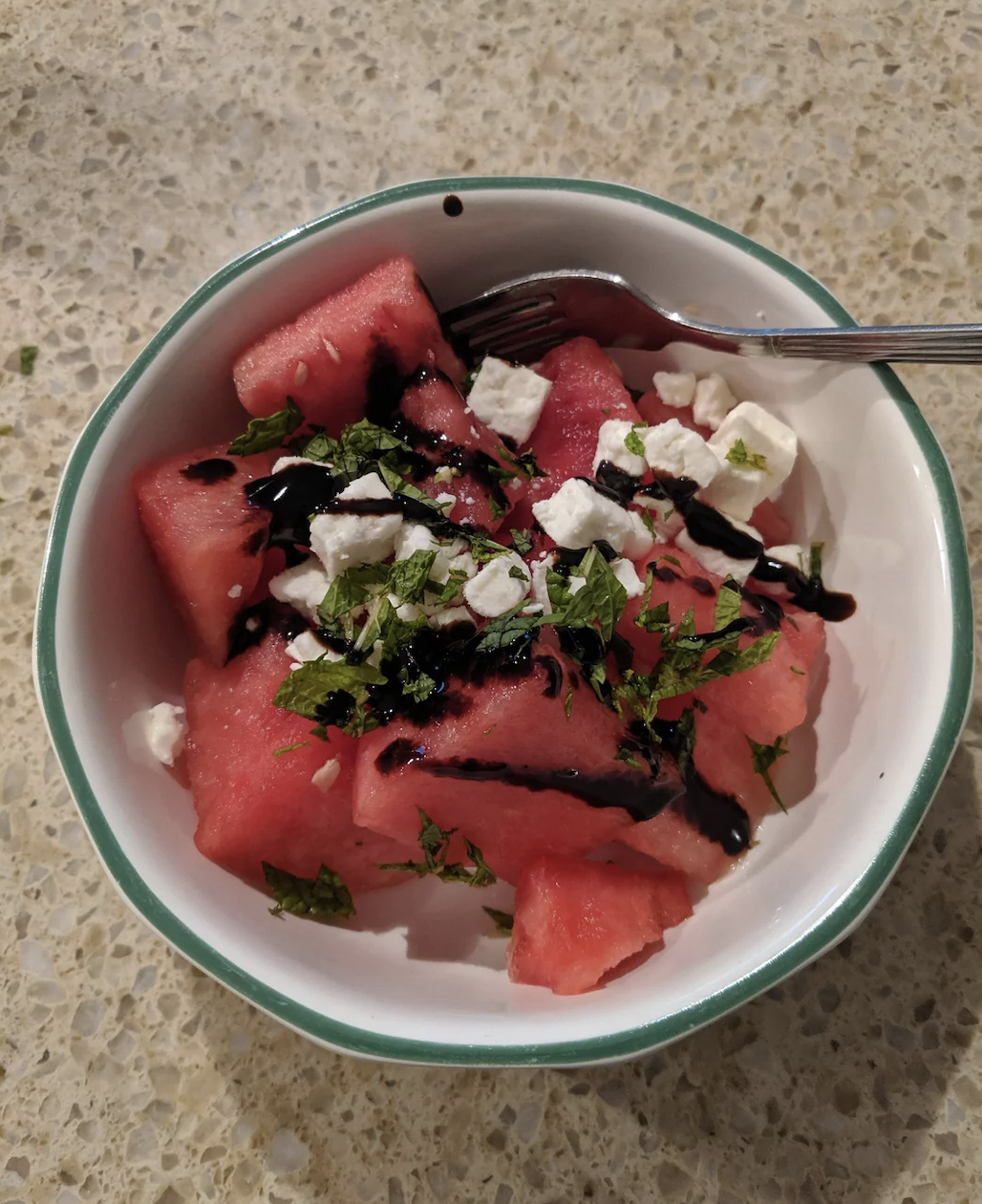 Bowl of watermelon cubes topped with feta cheese, chopped herbs, and a drizzle of balsamic glaze, with a fork on the side