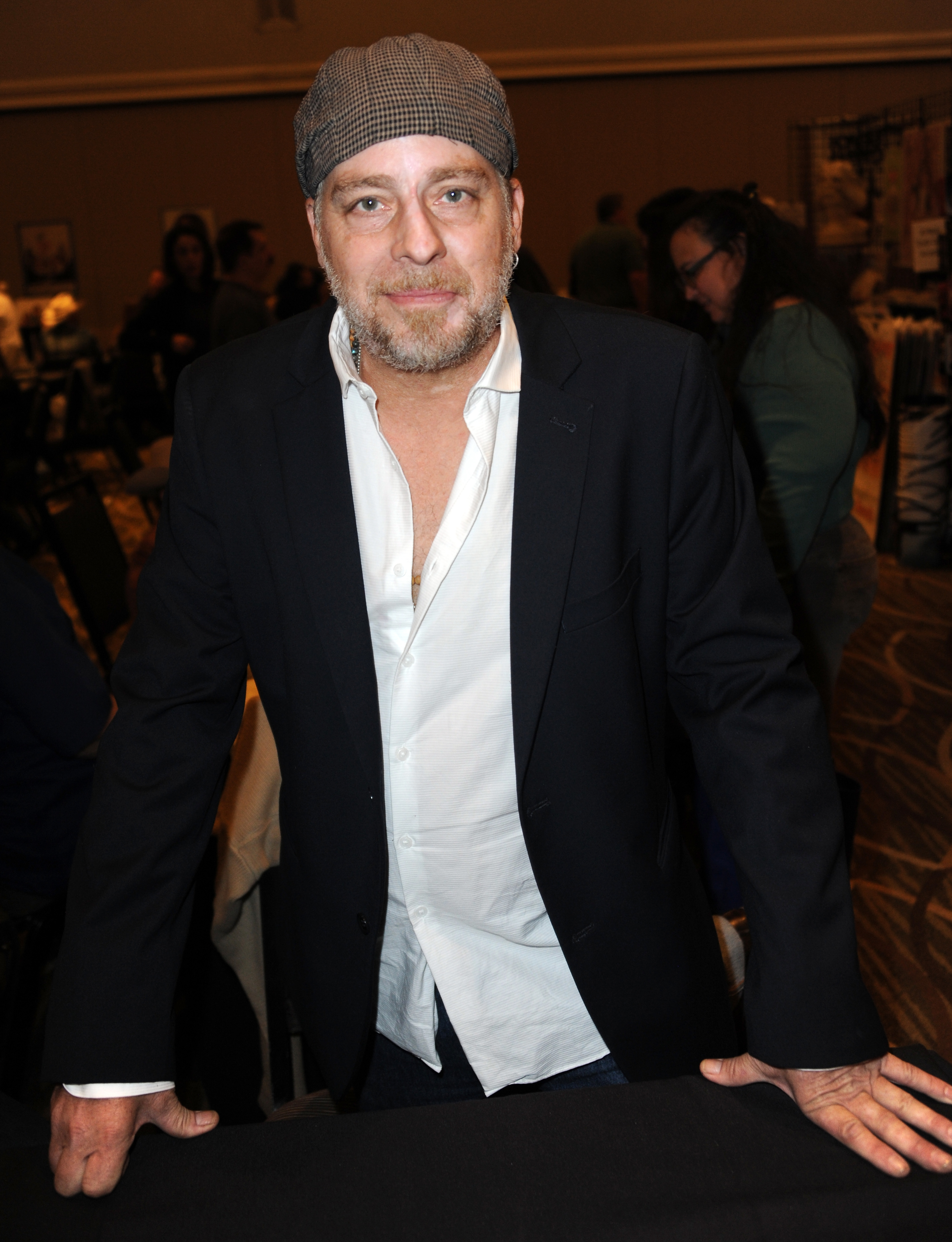 A man stands leaning against a black table, wearing a white shirt under a dark blazer and a cap