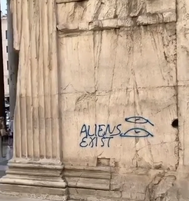 Graffiti on an ancient stone building reads "ALIENS EXIST" with two UFO drawings