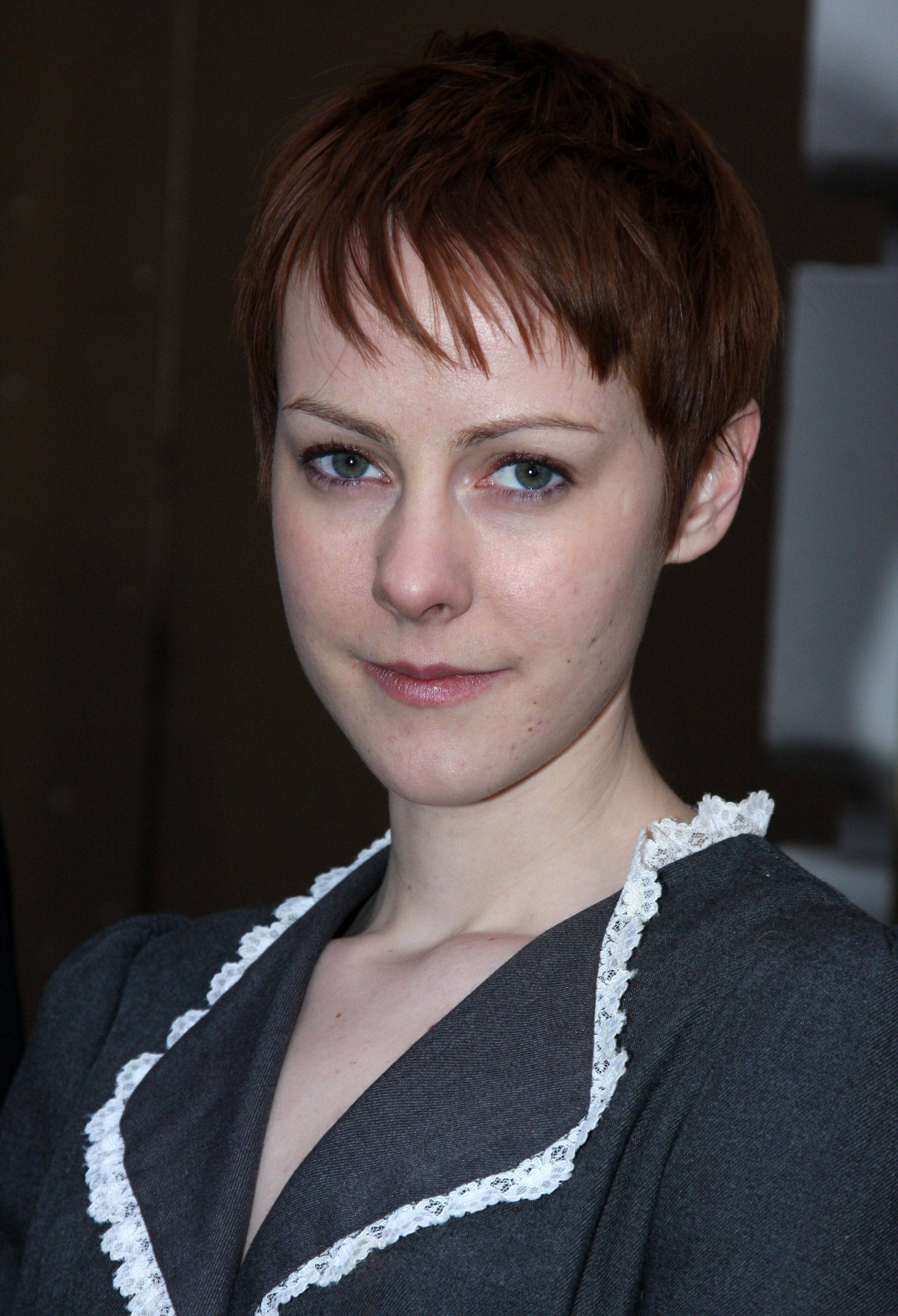 A woman with short brown hair and a neutral expression is wearing a dark dress with white lace trim on the collar