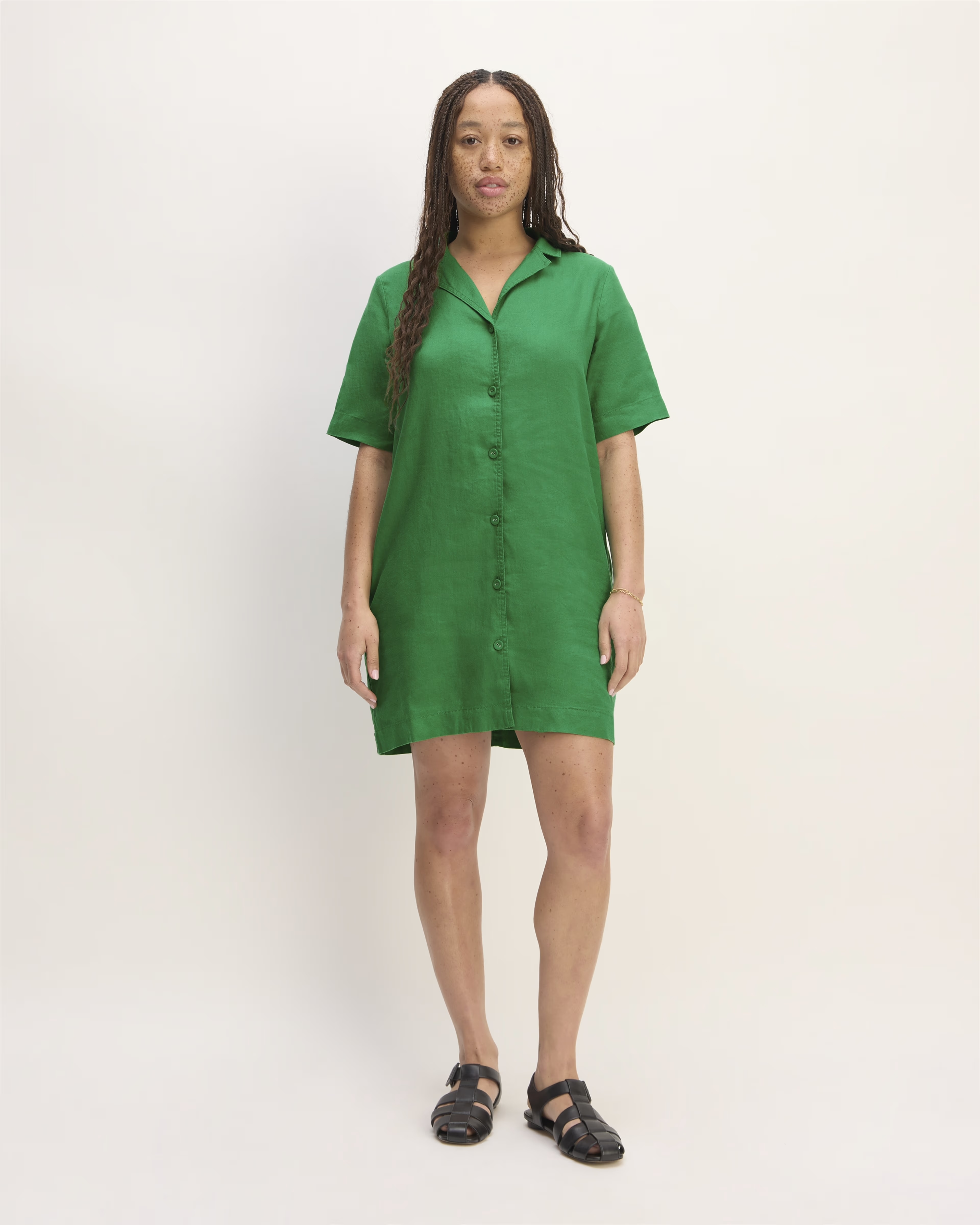 A woman wearing a short-sleeve, green button-down dress and black sandals stands against a plain background