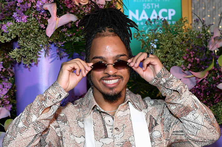 Man with dreadlocks, smiling while adjusting his sunglasses, in front of floral backdrop at the 55th NAACP event