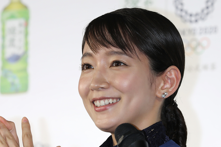 女性がイベントで話しています。背景にはオリンピックのロゴと緑茶のボトルが写っています。