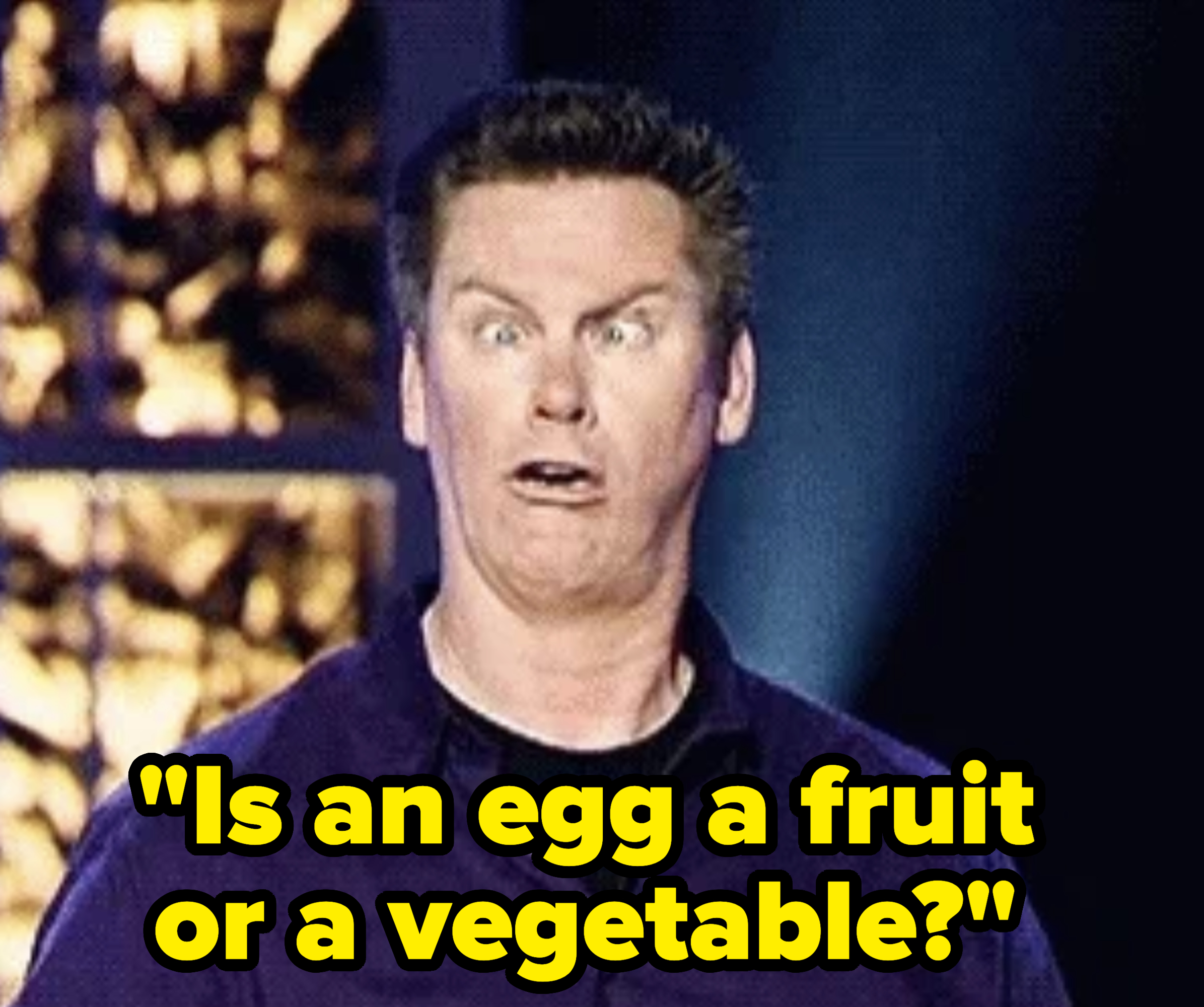 A man with a surprised and exaggerated facial expression, wearing a button-up shirt, stands on a stage