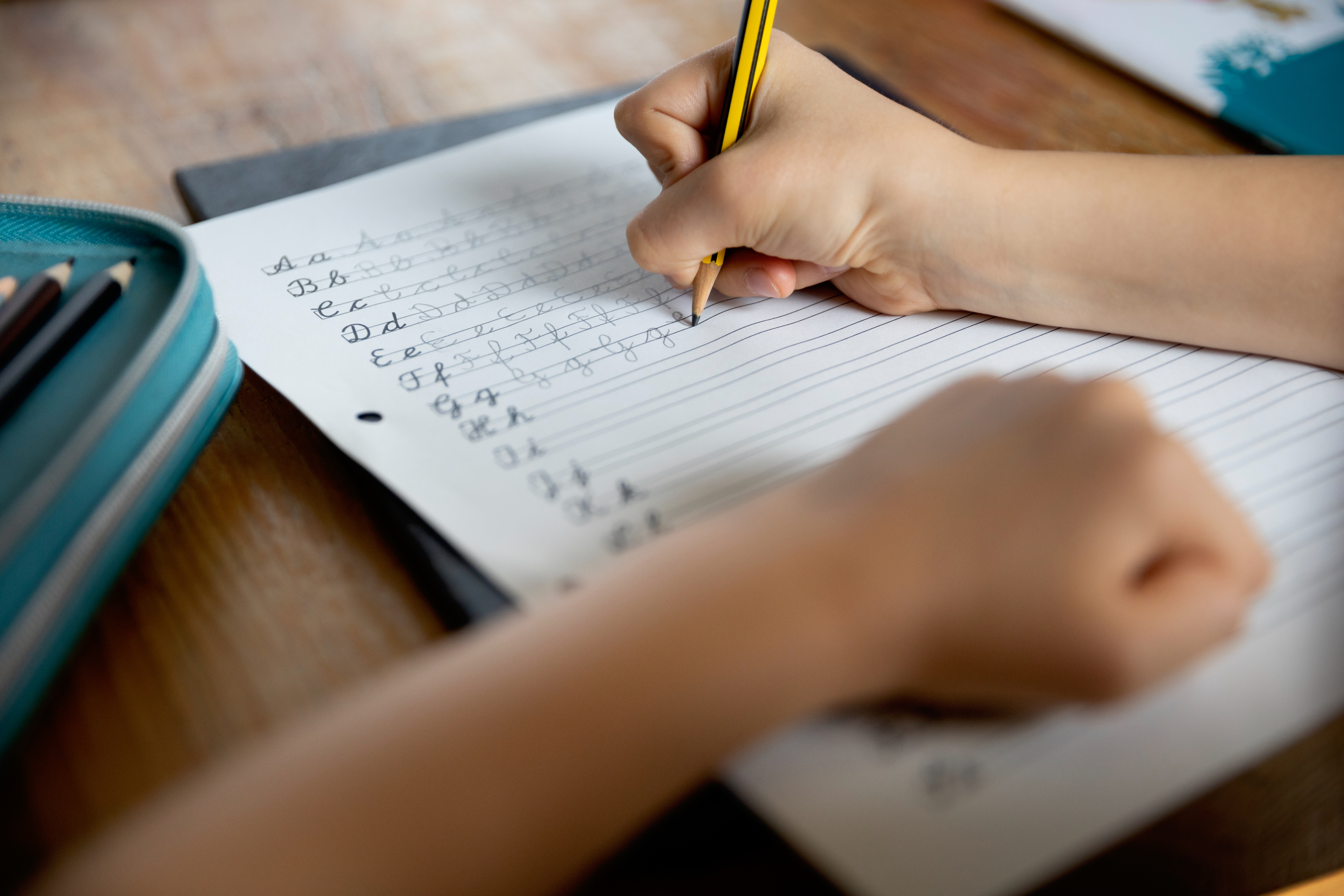 A child practices cursive writing on lined paper with a pencil, while resting their other arm on the desk